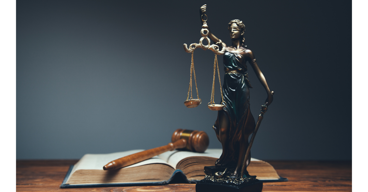 Une petite statue de Thémis, déesse grecque de la justice, tenant une balance et un glaive, et ayant les yeux bandés, est posée sur une table en bois devant un livre ouvert et un marteau de juge.